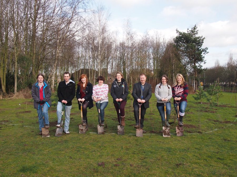A group of male and female students stand in a line in an open field. They pose with shovels as if about to break ground.